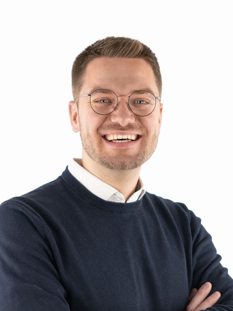 Porträt von David Jahn: Ein freundlich blickender Mann mit Brille, kurzen braunen Haaren und Bart. Er trägt einen dunkelblauen Pullover über einem weißen Hemd und hat die Arme verschränkt. Der Hintergrund ist weiß.