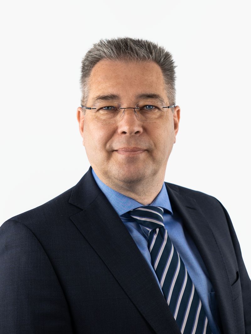 Porträt von Rechtsanwalt Johannes Kuhn: ein Mann mittleren Alters mit Brille, kurzem grauem Haar, im dunklen Anzug mit blauem Hemd und gestreifter Krawatte. Er blickt professionell und freundlich.