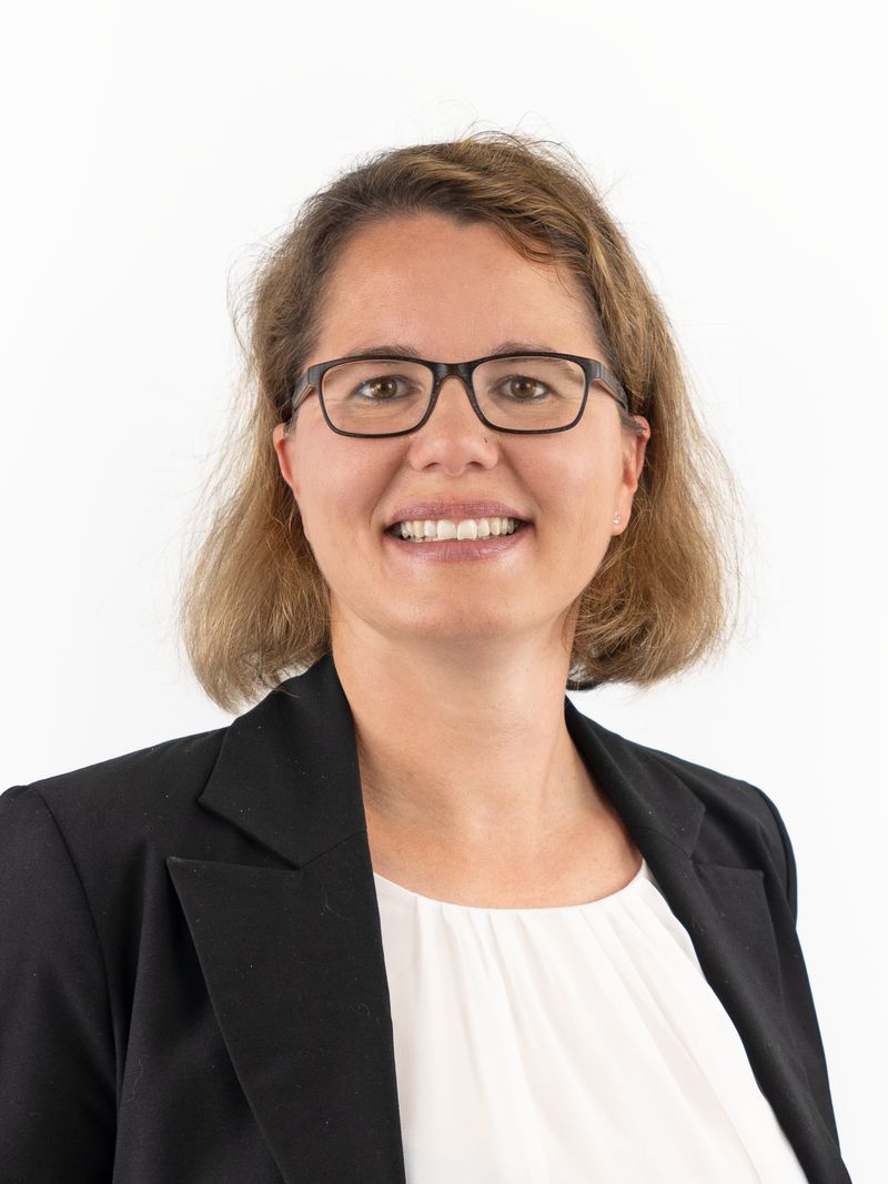 Porträt von Rechtsanwältin Katharina Benedikt. Sie hat schulterlange, braune Haare und trägt eine Brille. Sie lächelt und trägt einen schwarzen Blazer über einer weißen Bluse.