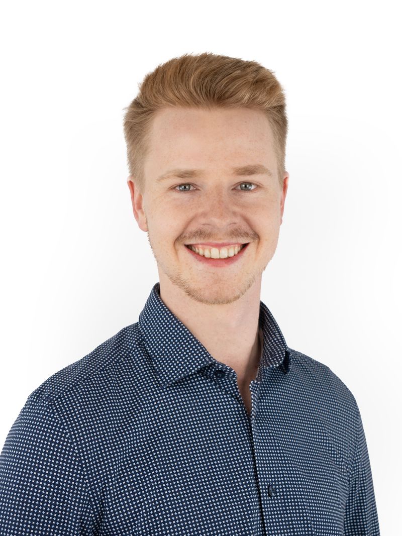 Porträt von Kieran Schreck: ein freundlich lächelnder junger Mann mit kurzen blonden Haaren, hellem Bartansatz und dunkelblau gemustertem Hemd vor weißem Hintergrund.