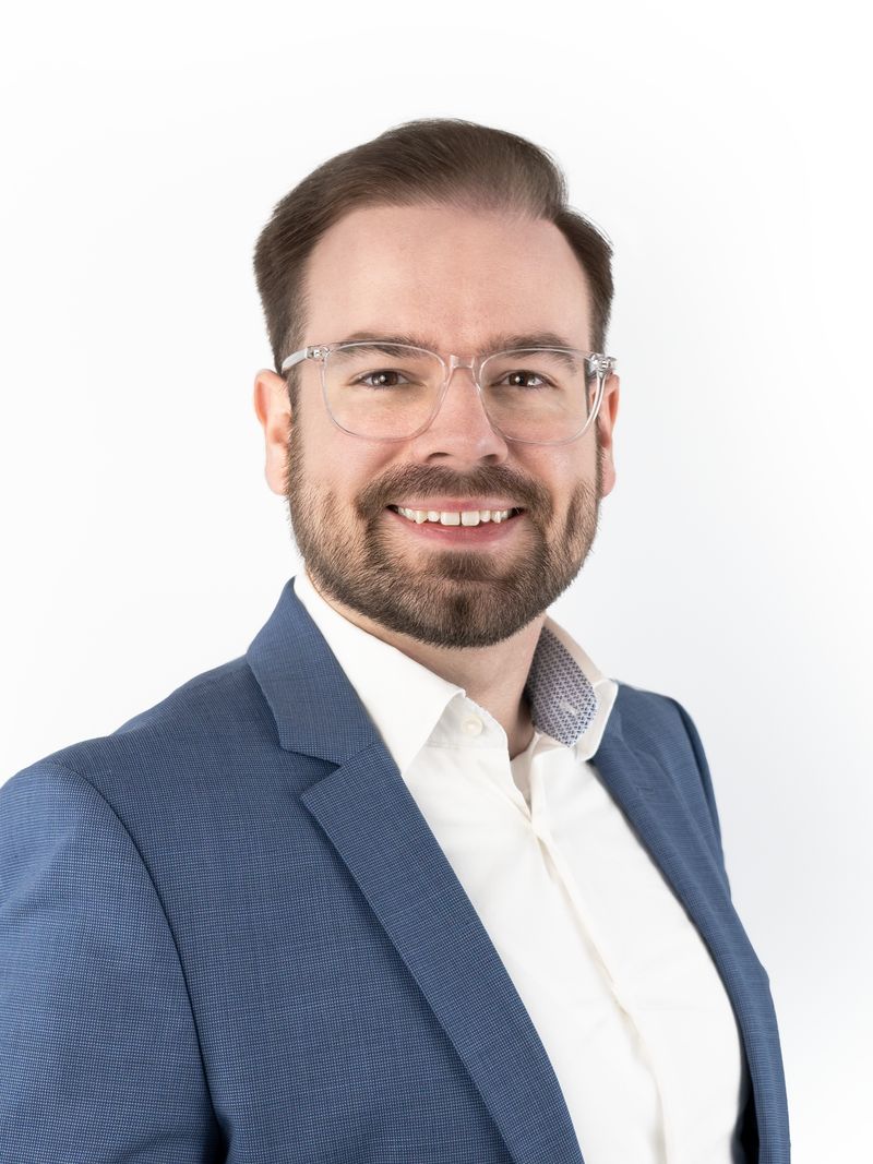 Porträt von Marlon Lazar: freundlich lächelnder Mann mit transparenter Brille, dunklem Bart, weißem Hemd und blauem Sakko vor hellem Hintergrund.