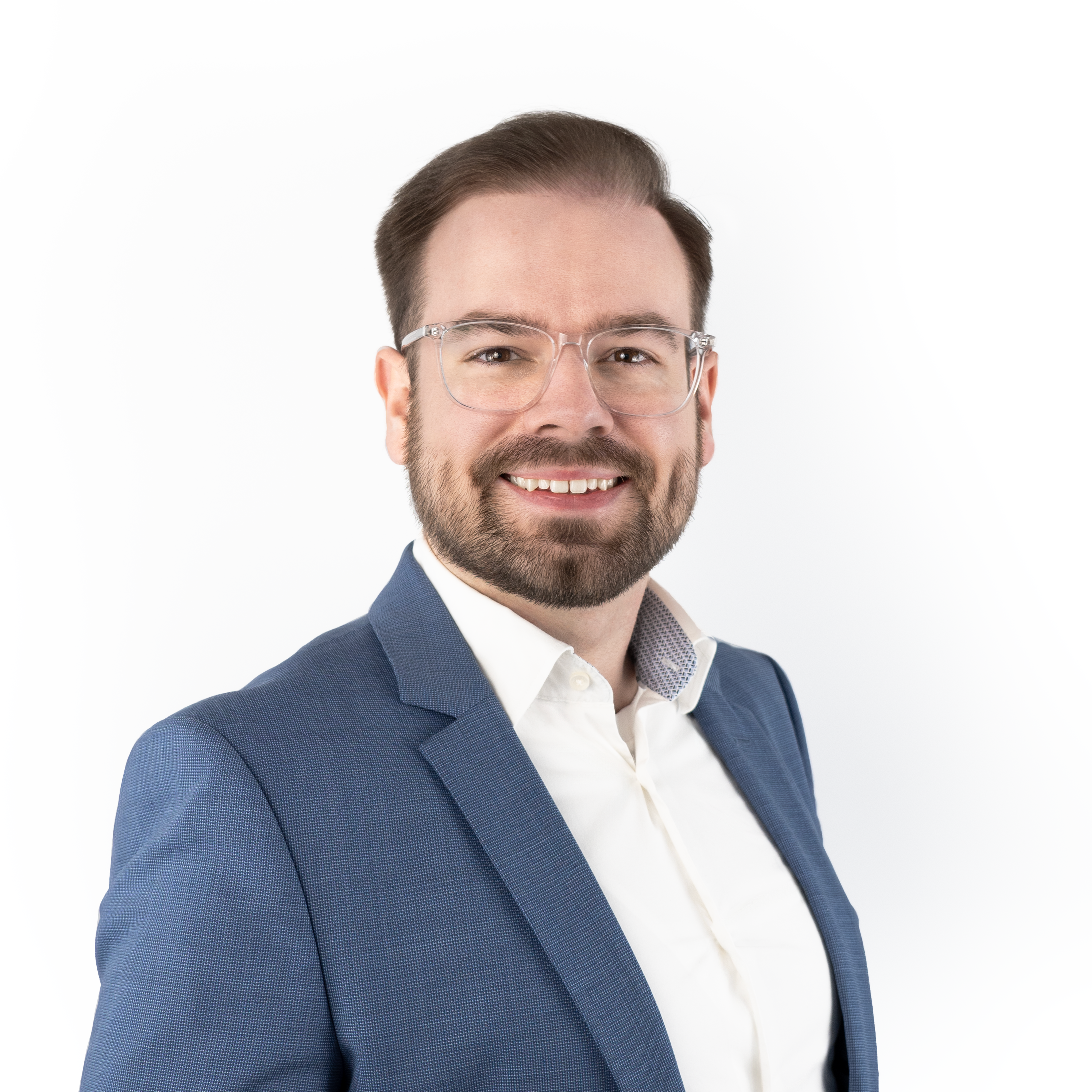 Porträt von Marlon Lazar: freundlich lächelnder Mann mit transparenter Brille, dunklem Bart, weißem Hemd und blauem Sakko vor hellem Hintergrund.